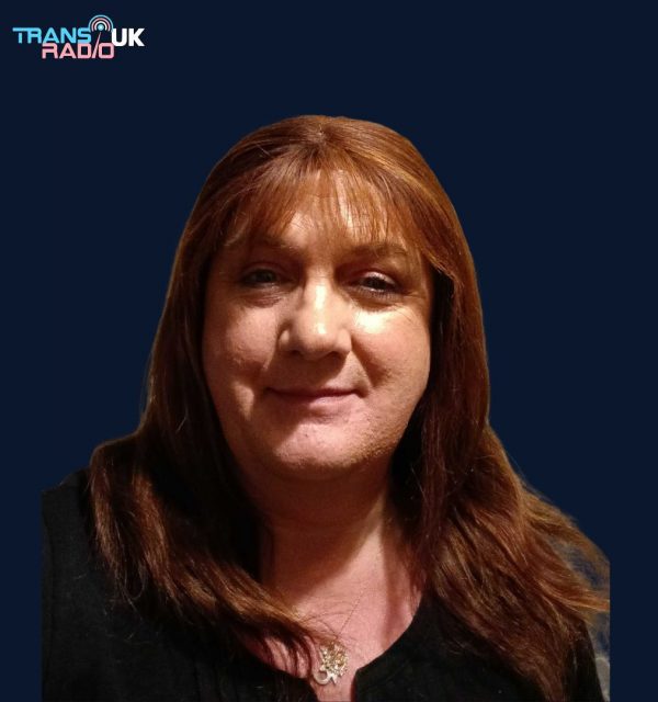 Andrea, presenter at Trans Radio UK, with long auburn hair and a warm smile, wearing a black top and necklace against a dark background with the Trans Radio UK logo.